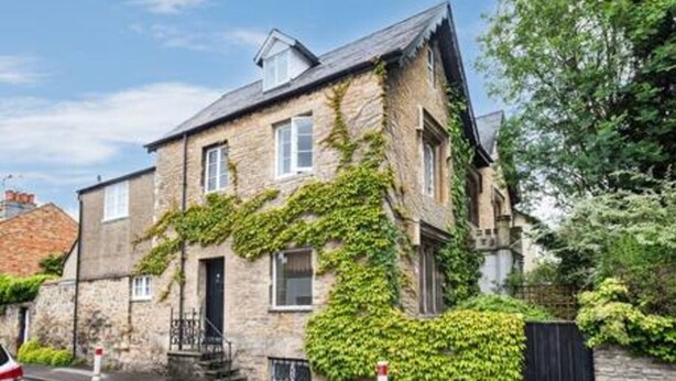 Une maison en pierre, recouverte de lierre, avec une porte d’entrée bien visible et un petit jardin devant.