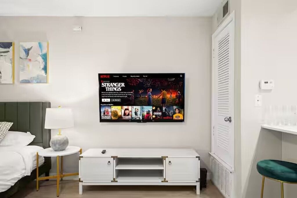 A modern hotel room with a bed, a bedside table with a lamp, a white TV stand, and a wall-mounted TV displaying the 'Stranger Things' series.
