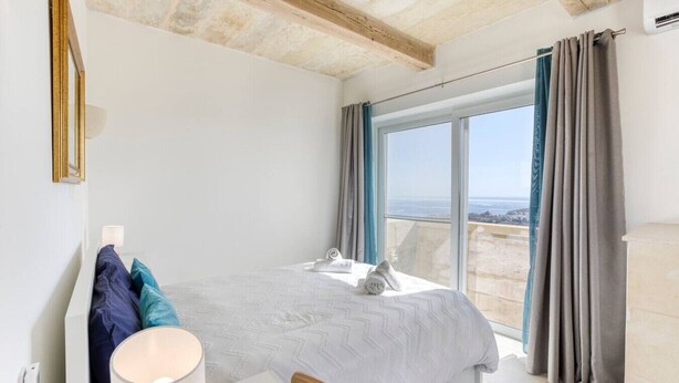 Una camera da letto con un letto, un comodino, una lampada e vista mare attraverso ampie finestre.