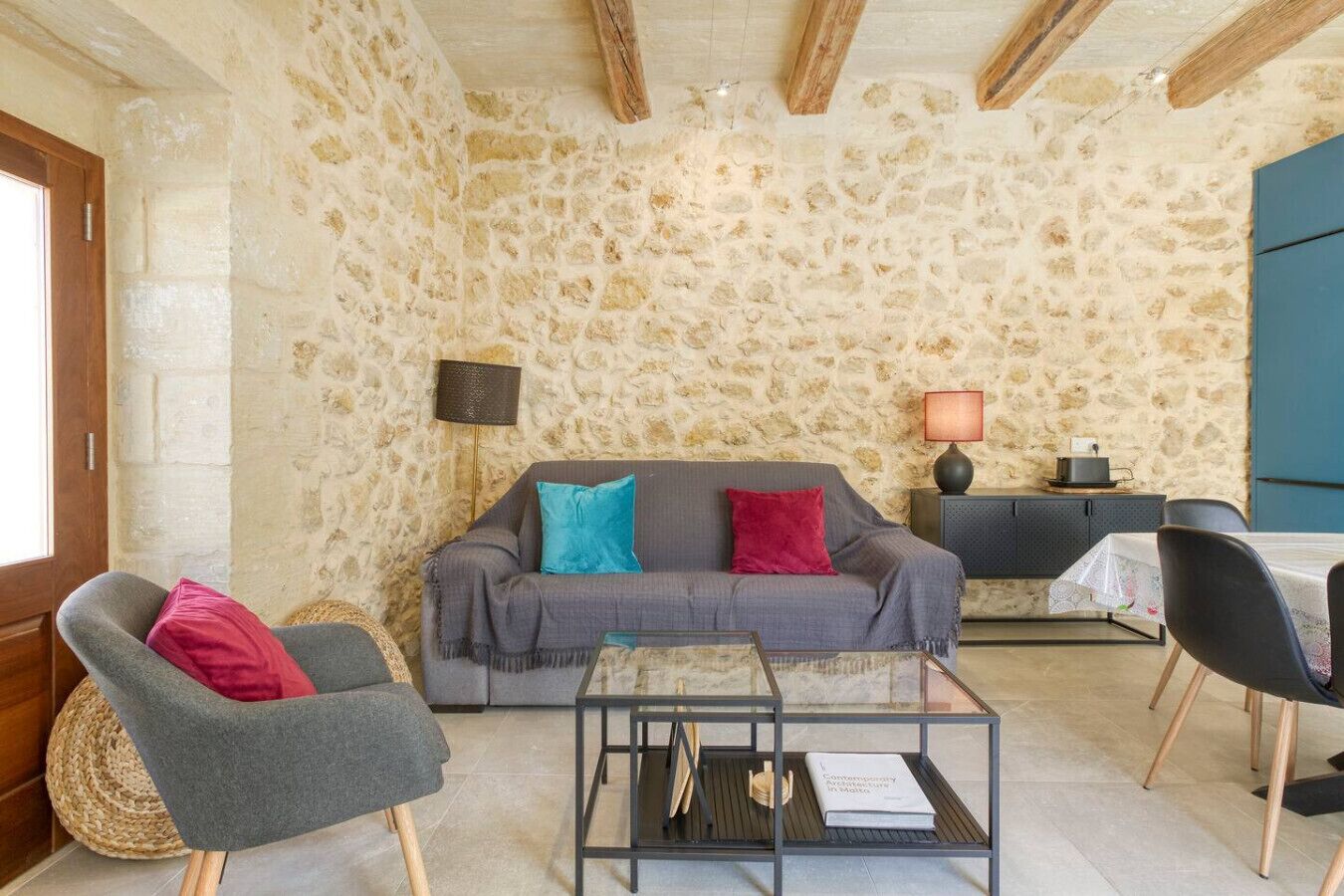 A cozy living room with a stone wall, a grey sofa with colorful cushions, a glass coffee table, and a dining area with a table and chairs.
