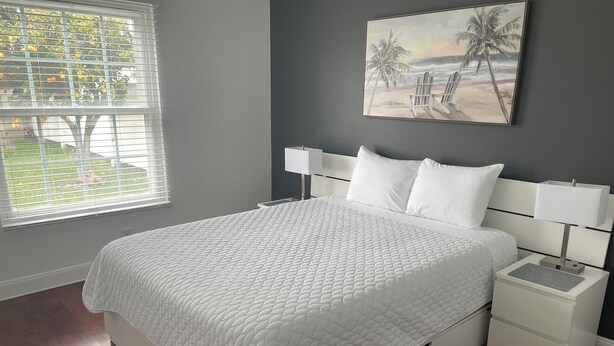Quarto com cama, mesas de cabeceira brancas, pintura de uma praia e janela com persianas.