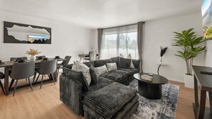 Ett modernt vardagsrum med en hörnsoffa, ett matbord och stolar. Det finns ett stort fönster med gardiner, en tavla på väggen och en krukväxt.