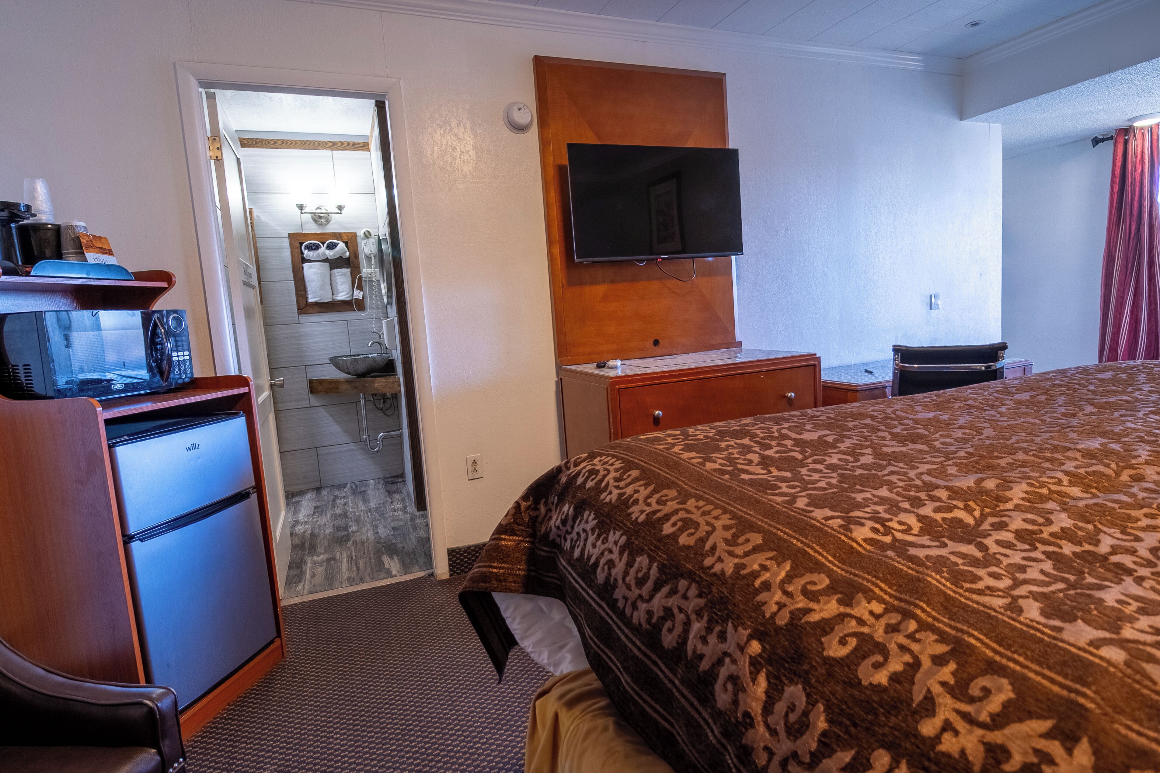 A hotel room with a bed, a desk, a television, a minibar, and a bathroom visible through an open door.