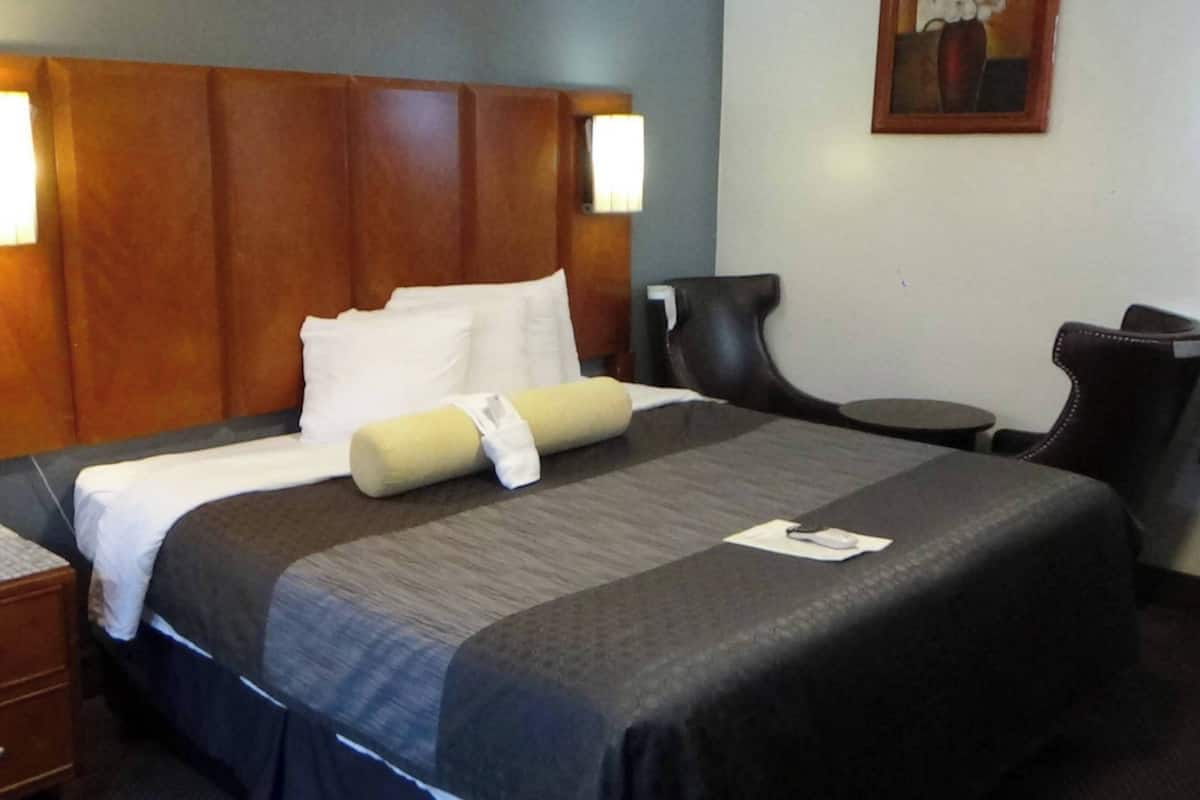 Une chambre d’hôtel avec une tête de lit en bois, un lit avec une literie blanche et beige, une petite table de chevet avec une lampe, une chaise et un bureau avec une autre chaise.