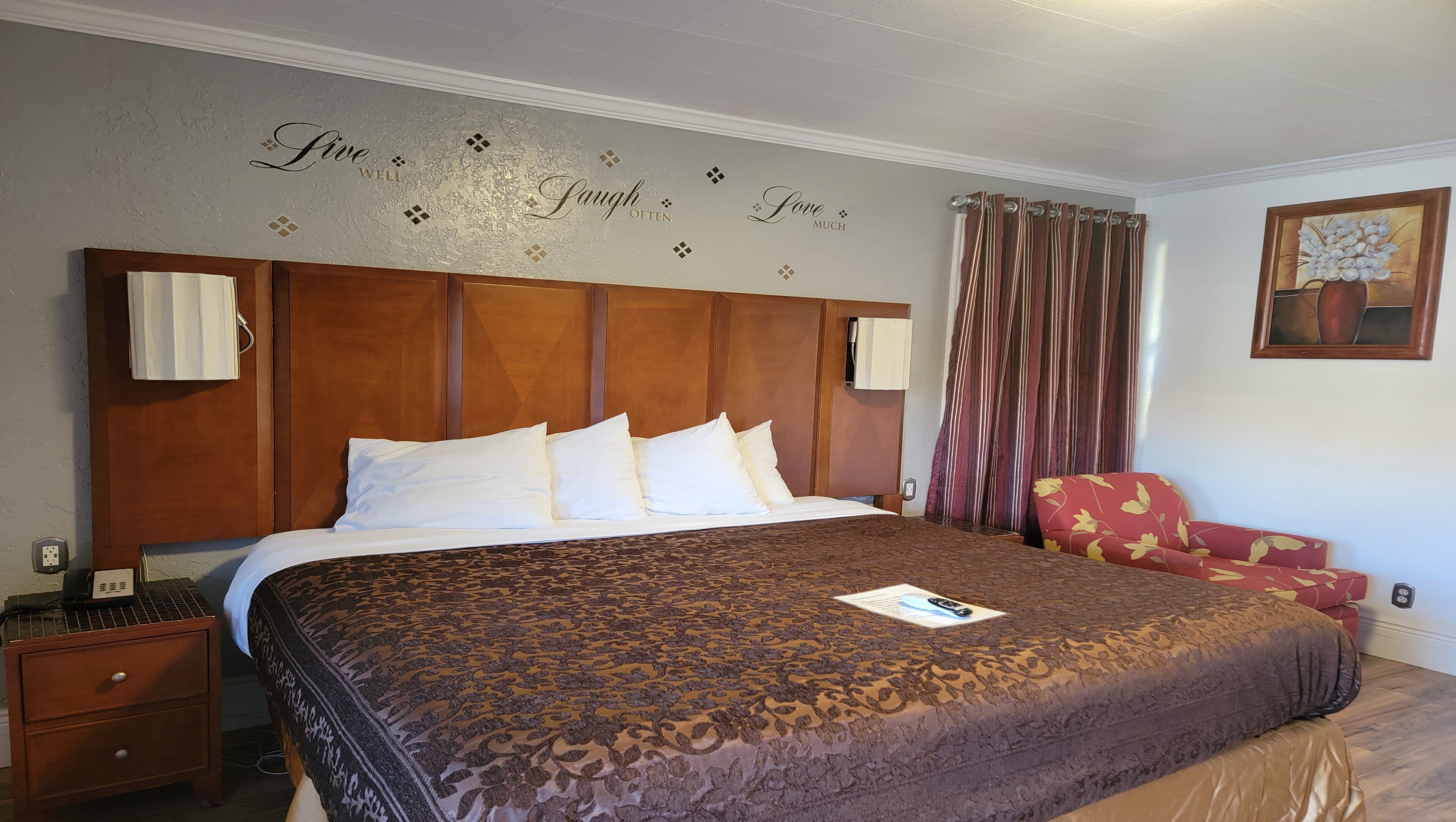 Habitación de hotel con una cama grande, cabecera de madera, un sillón rojo con estampado floral, un cuadro en la pared y una frase decorativa.