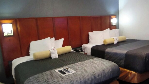 Habitación de hotel con dos camas, cada una con una almohada y una manta. Hay un control remoto en el suelo entre las camas.