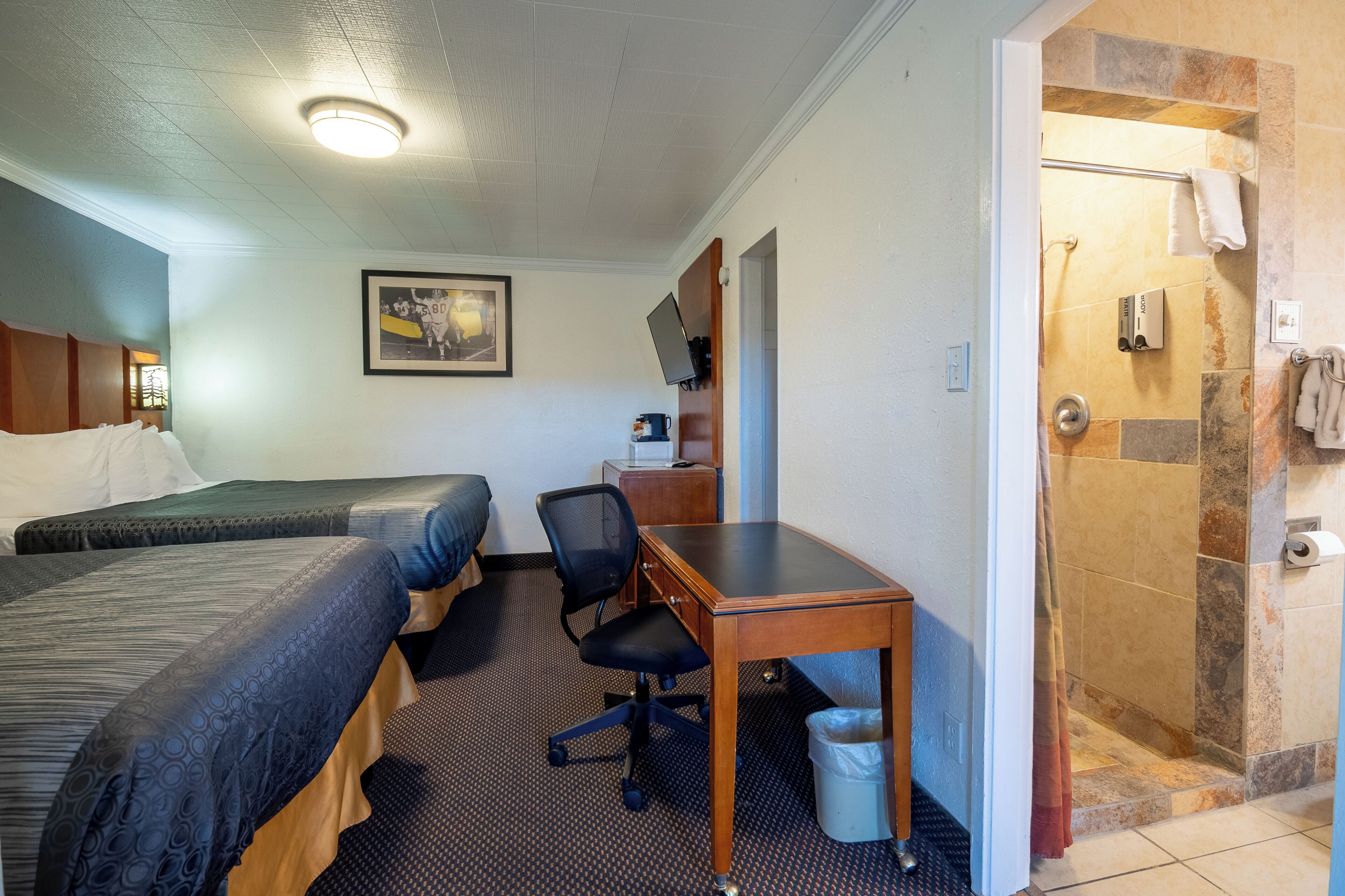 Een hotelkamer met twee bedden, een bureau, een stoel, een douche en een ingelijst schilderij aan de muur.