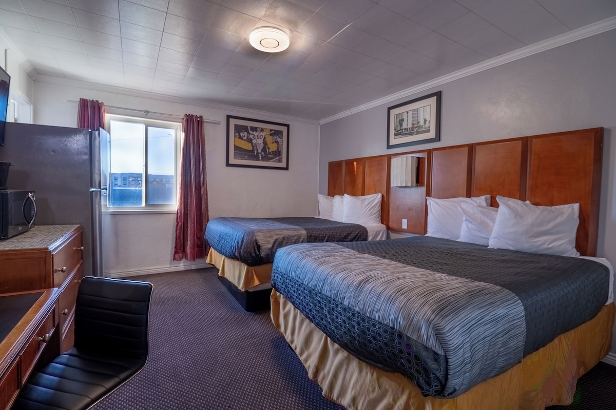 A hotel room with two beds, a desk, a chair, a window with curtains, and a framed picture on the wall.