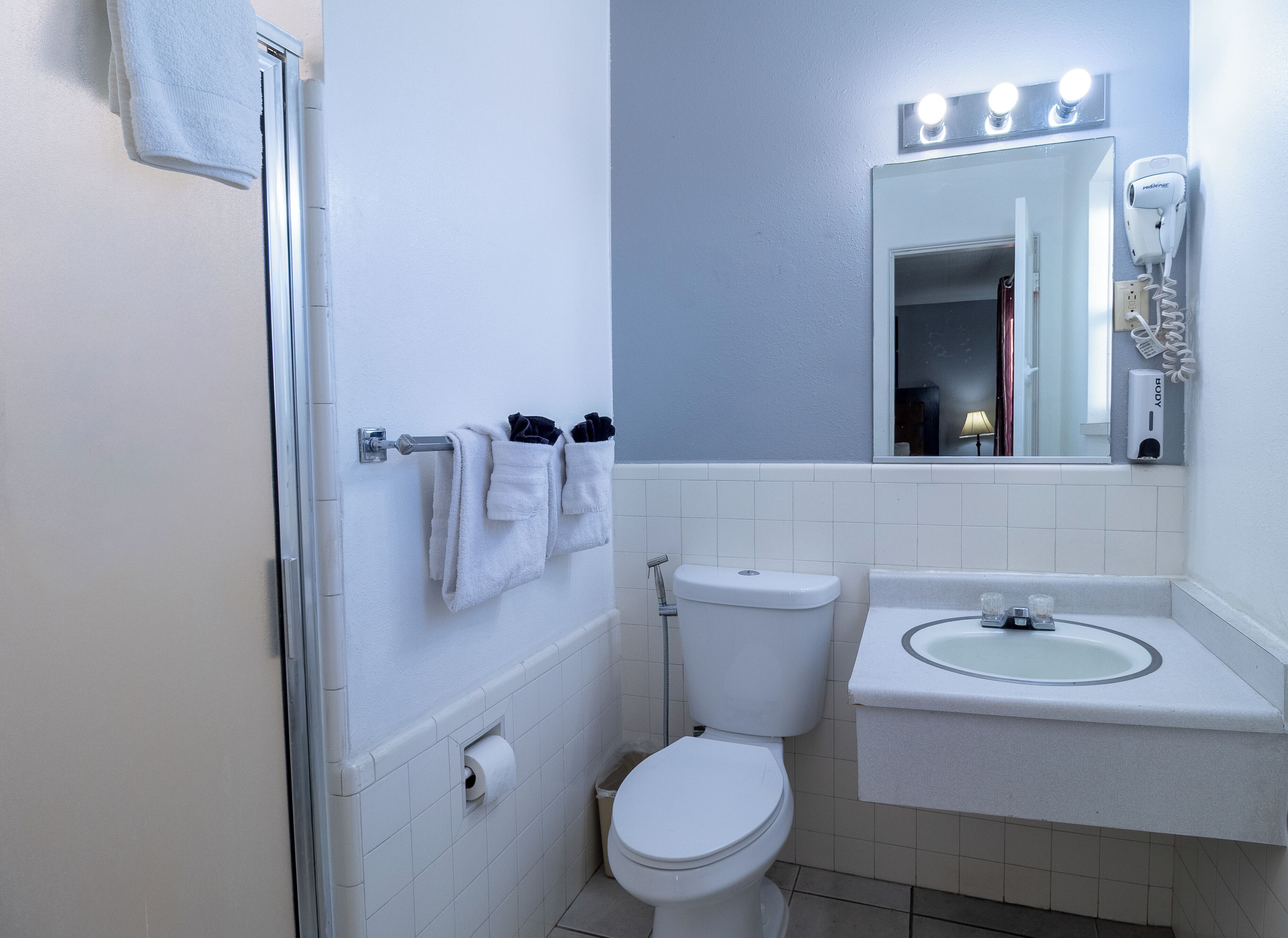 Un baño con inodoro, lavamanos, toallero y un espejo con luces arriba.