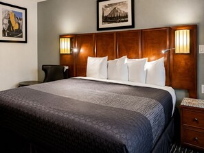 A hotel room with a large bed, a wooden headboard, two wall-mounted lamps, a framed picture above the bed, and a small table with a chair.