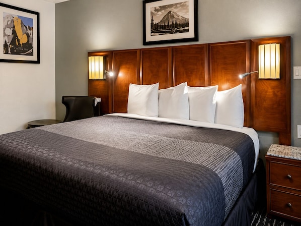 A hotel room with a large bed, a wooden headboard, two wall-mounted lamps, a framed picture above the bed, and a small table with a chair.