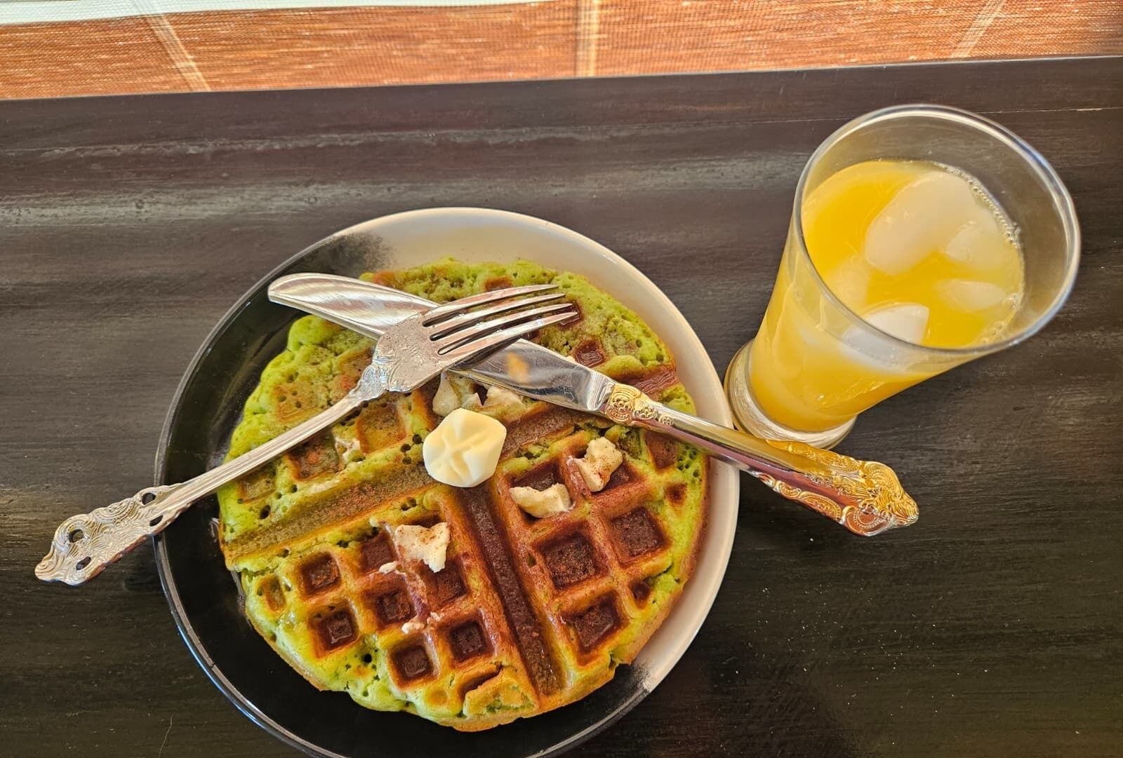 A plate of green waffles with butter and a glass of orange juice on a dark wooden table.
