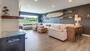 Un salon moderne avec un canapé, des poufs et une table basse. Un surf est accroché au mur, et un petit coin bar avec des tabourets est aménagé.