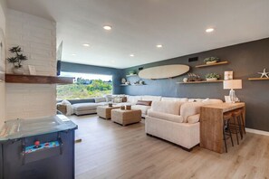 Un salon moderne avec un canapé, des poufs et une table basse. Un surf est accroché au mur, et un petit coin bar avec des tabourets est aménagé.