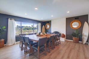Un coin repas avec une grande table et des chaises, une vue par la fenêtre et une planche de surf accrochée au mur.