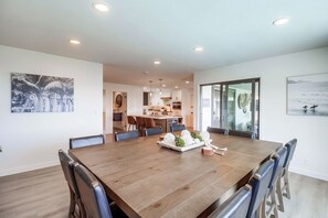 A modern dining area with a wooden table and chairs, a kitchen in the background, and artwork on the walls.