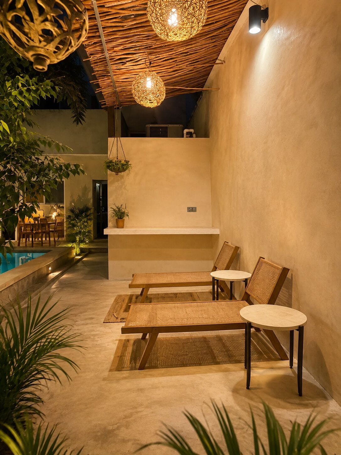 An indoor pool area with lounge chairs and a small table, surrounded by plants and a ceiling with woven light fixtures.