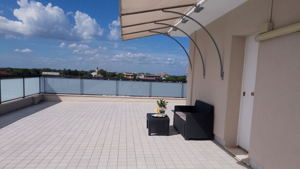 Een terras op het dak met betegelde vloer, een overdekte zithoek met een tafel en stoelen, en een uitzicht op een wateroppervlak en gebouwen in de verte.
