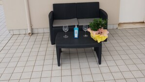 Een zwarte rieten bank met een kussen, een zwarte tafel met twee wijn glazen en een fles, en een potplant met gele bloemen.