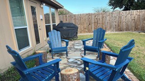 Une terrasse avec quatre chaises Adirondack bleues disposées autour d’une table en pierre centrale, une clôture en bois en arrière-plan et un barbecue.