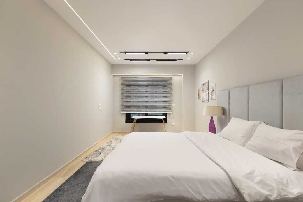 A modern hotel room with a large bed, a bedside table, a lamp, and a window with blinds.