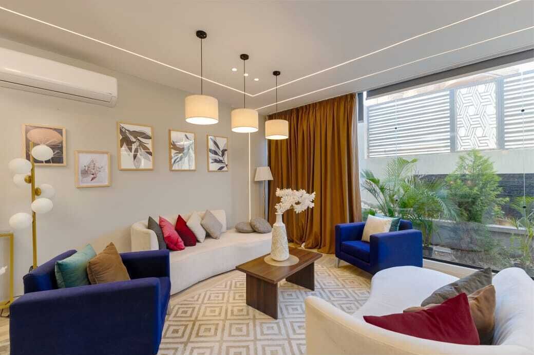 A modern living room with a white sofa, blue armchairs, and a coffee table. There are framed artworks on the wall and a large window with curtains.