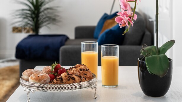 A breakfast setup with pastries, fruit, and orange juice on a tray, a potted orchid, and a modern living room in the background. - Stylish 2 Bed Apartment Central Brighton, The Lanes Sleeps 8 (Brighton and Hove)