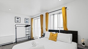 A hotel room with a large bed, a black headboard, and yellow curtains. There is a black metal rack with towels and a small potted plant on the side table. - Stylish 2 Bed Apartment Central Brighton, The Lanes Sleeps 8 (Brighton and Hove)