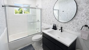 Ein modernes Badezimmer mit einer gläsernen Duschkabine, einer weißen Toilette, einem Waschtisch mit schwarzem Waschbecken und einem runden Spiegel.