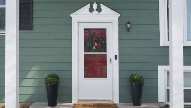 Ein grünes Haus mit weißem Vorbau, einer rot umrandeten Tür und je einem Blumentopf auf beiden Seiten.