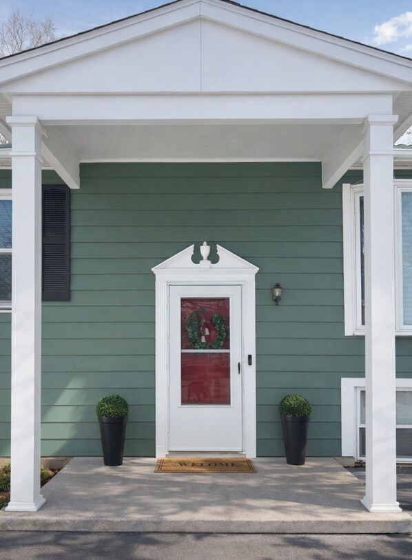 A green house with a white porch, a red-framed door, and two potted plants on either side.