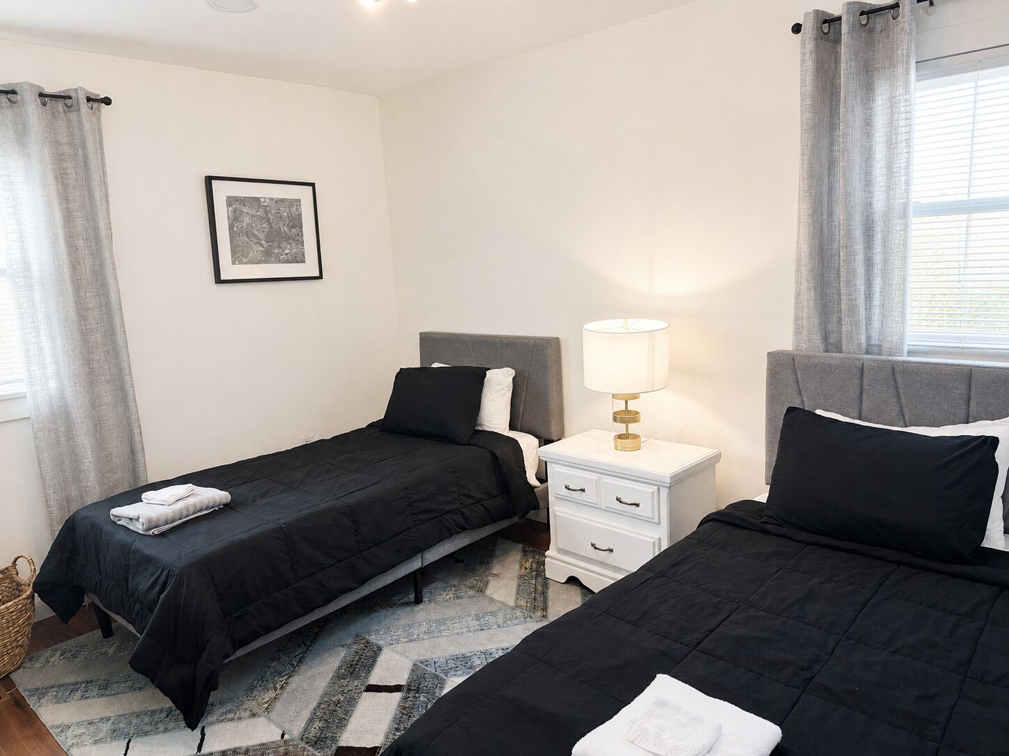 Una habitación de hotel con dos camas individuales, una mesita de noche con lámpara, un cuadro en la pared y una ventana con cortinas.