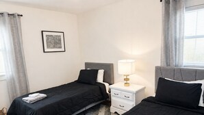 A hotel room with two single beds, a bedside table with a lamp, a framed picture on the wall, and a window with curtains. - Your Cortland Base Near SUNY (Cortland)