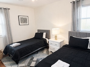 Una habitación de hotel con dos camas individuales, una mesita de noche con lámpara, un cuadro en la pared y una ventana con cortinas.