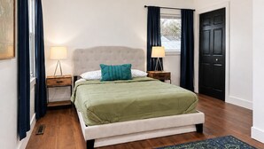 Ein Schlafzimmer mit einem Bett, Nachttischen, einer Lampe, einer Tür und einem Fenster mit Vorhängen.