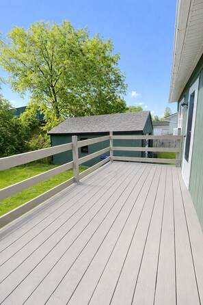 A wooden deck with a white railing, a green building in the background, and a tree.