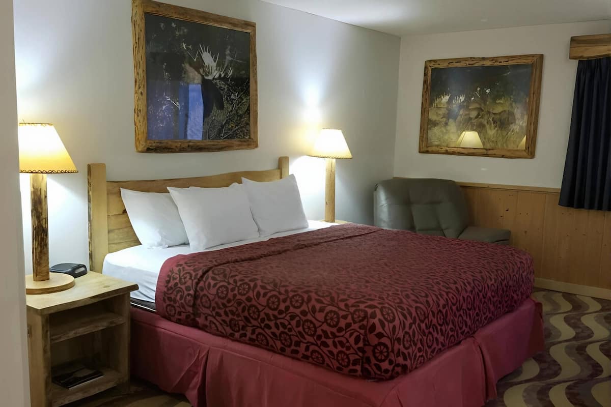 A hotel room with a large bed, two bedside lamps, a wooden headboard, a chair, and two paintings on the walls.