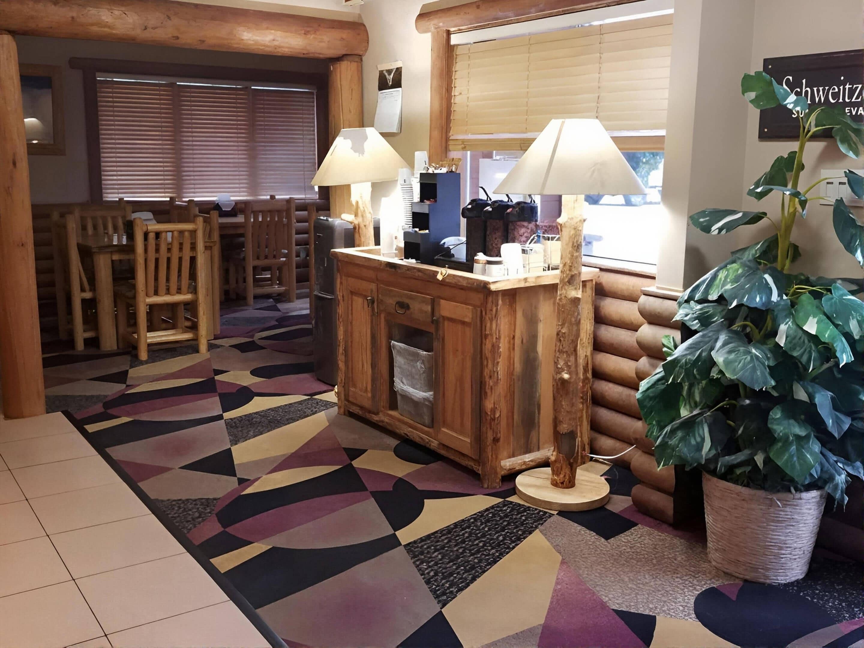 A cozy lobby with a wooden reception desk, two lamps, a large potted plant, and a patterned rug.