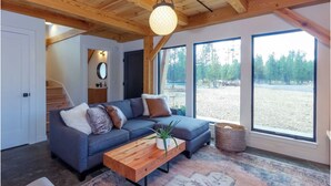 Un salon moderne avec un canapé d’angle gris, une table basse en bois et une grande fenêtre donnant sur une zone boisée.