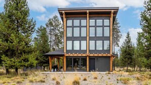 Un bâtiment moderne à deux étages, doté de grandes fenêtres et de poutres en bois, entouré d’arbres et bordé d’un sol en gravier.