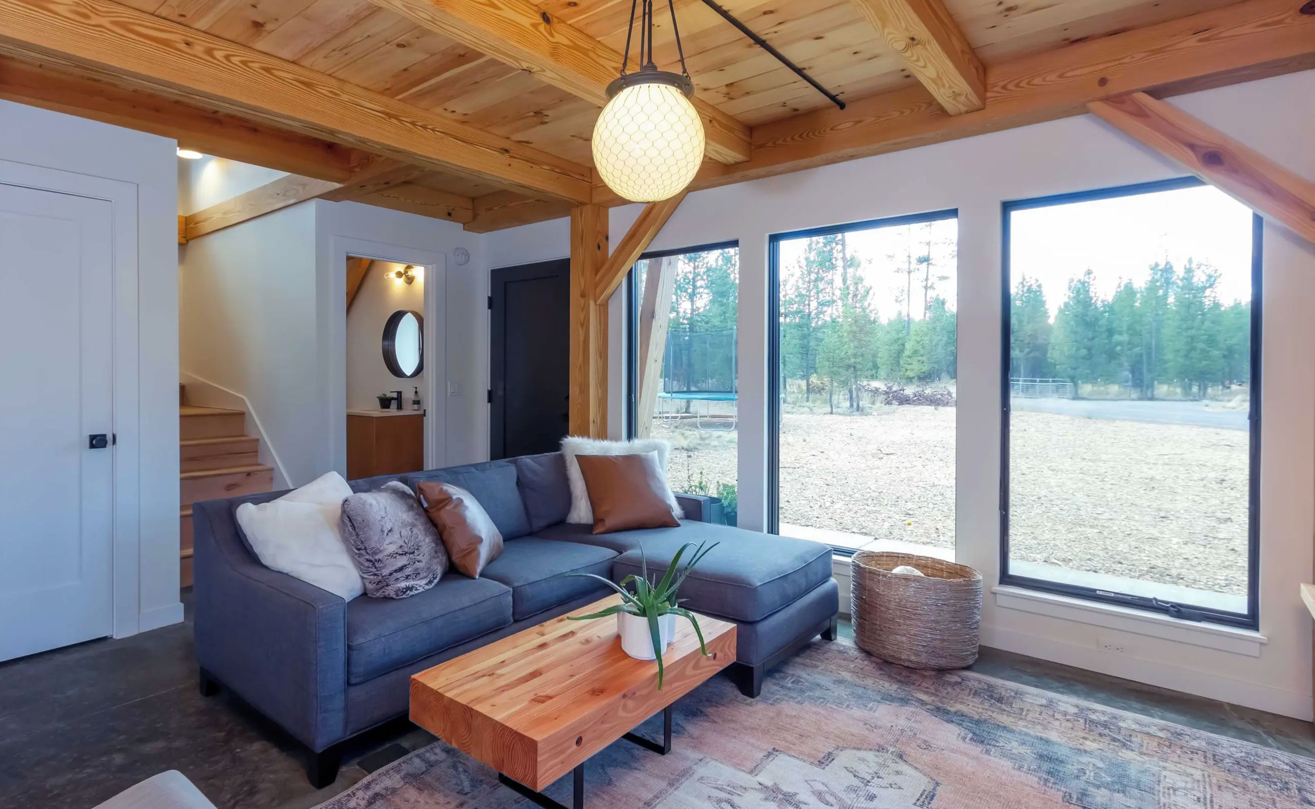 Een moderne woonkamer met een grijze hoekbank, een houten salontafel en een groot raam met uitzicht op een bebost gebied.