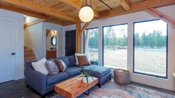 Een moderne woonkamer met een grijze hoekbank, een houten salontafel en een groot raam met uitzicht op een bebost gebied.