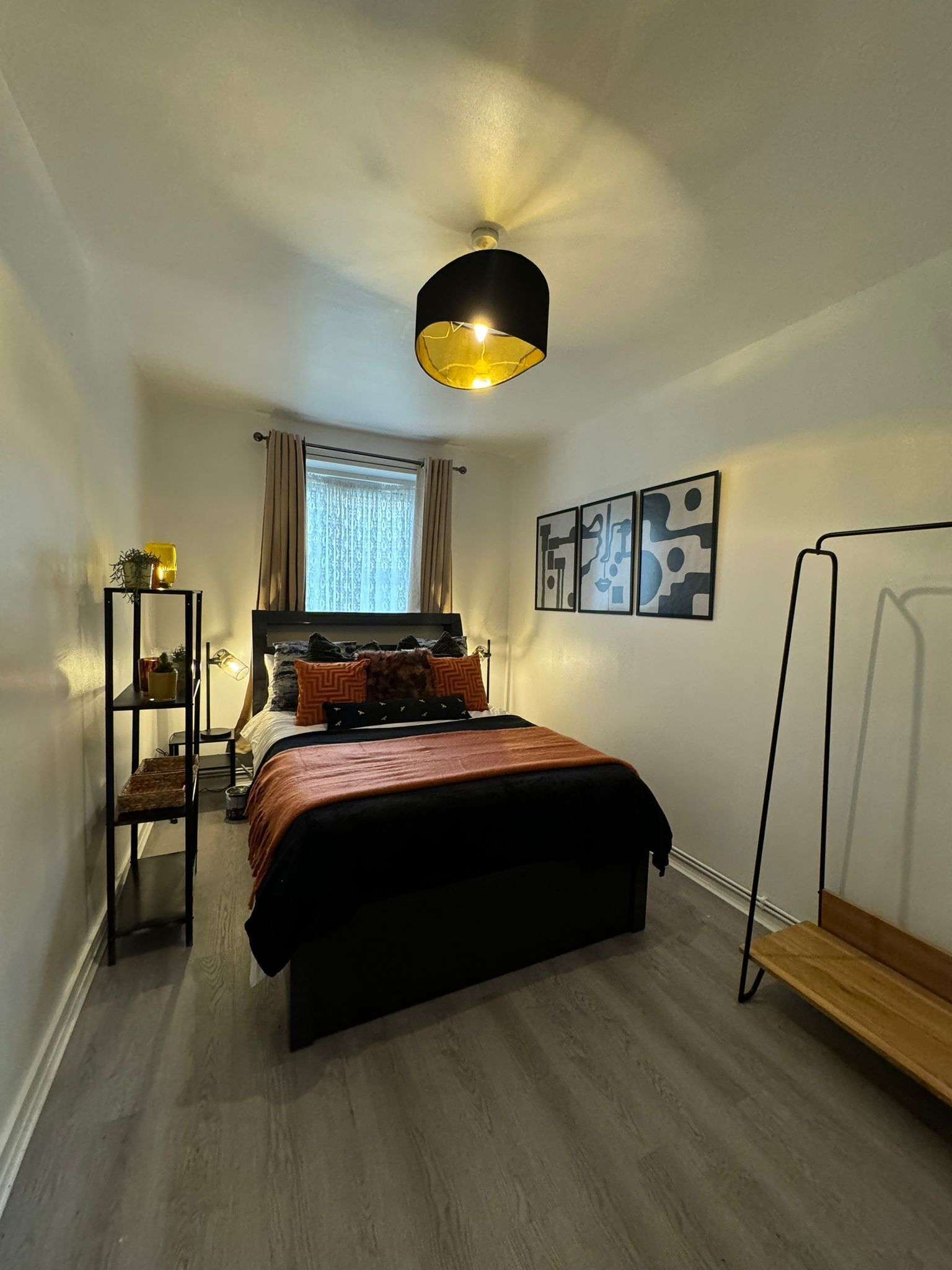 Ein Schlafzimmer mit Bett, Nachttisch, Regal, Fenster mit Vorhängen und Wandbildern.