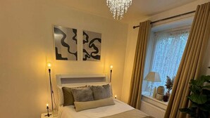 Ein Schlafzimmer mit Bett, Nachttischen, einer Stehlampe, einem Fenster mit Vorhängen und einer Deckenleuchte.
