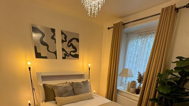 Ein Schlafzimmer mit einem großen Bett, Nachttischlampen, einem Kronleuchter, einem Fenster mit Vorhängen und einer Pflanze.