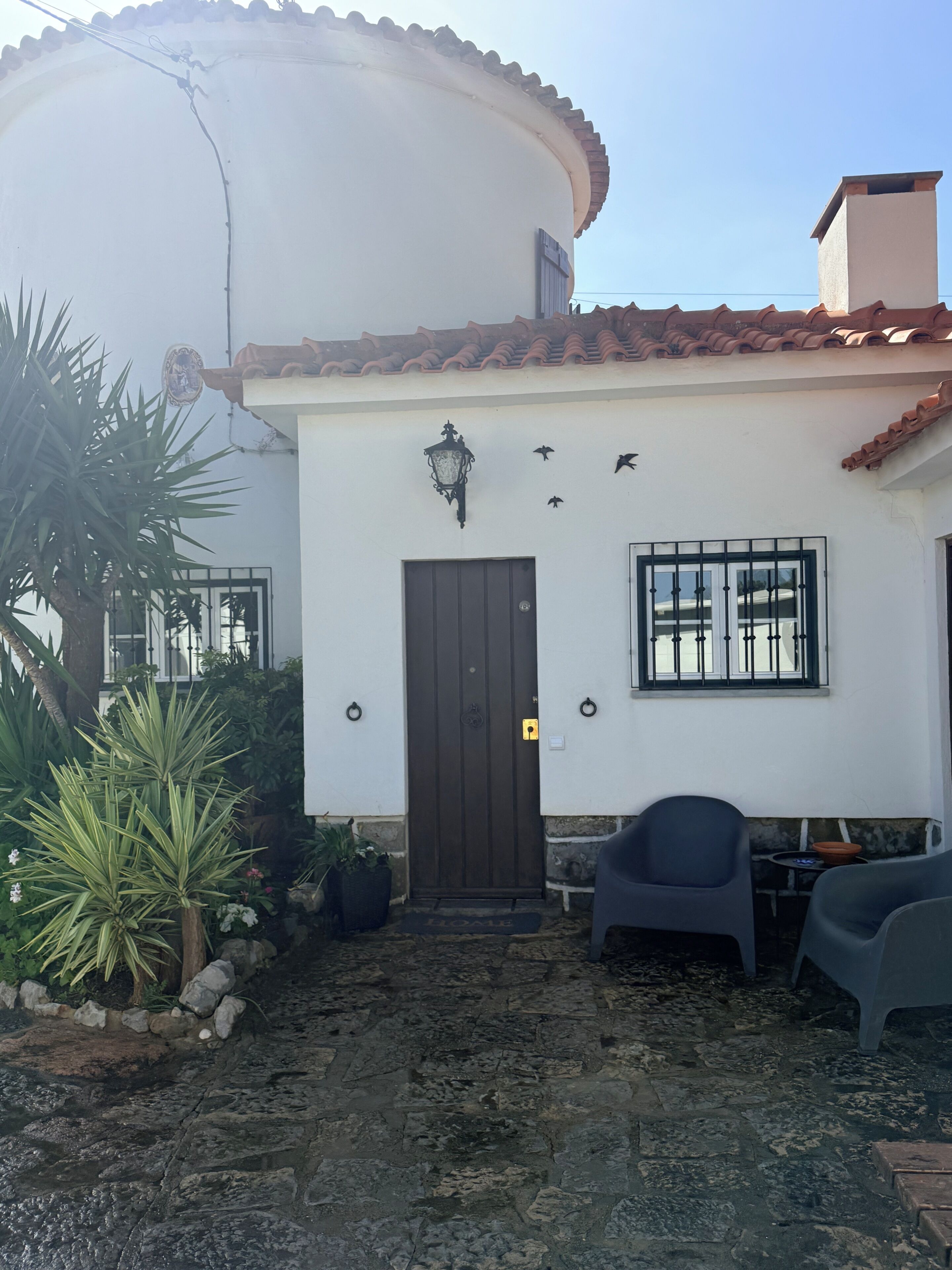 Un edificio blanco con una puerta marrón, una ventana con barrotes negros y un pequeño porche con sillas y plantas en macetas.