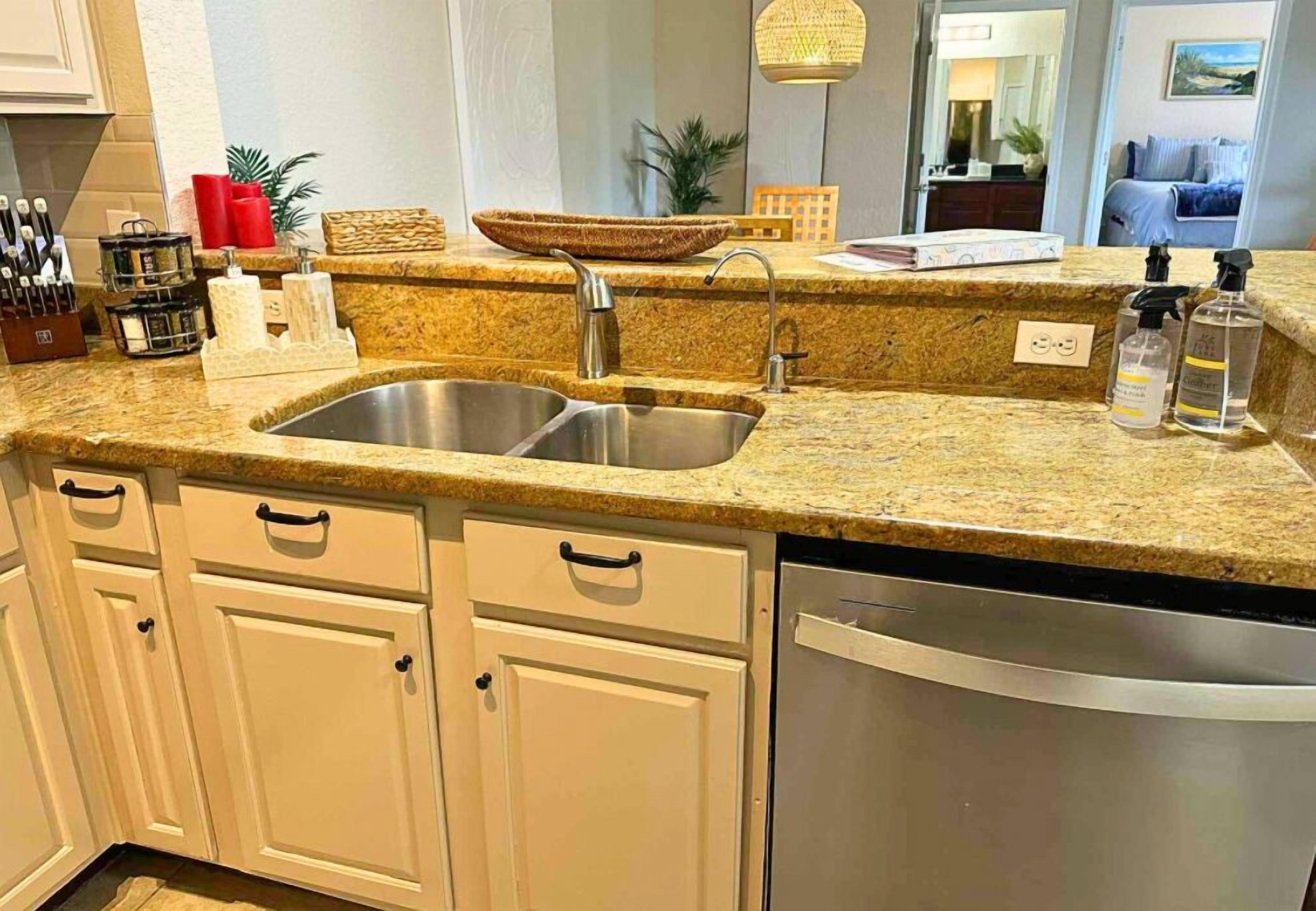 A kitchen with granite countertops, a stainless steel sink, and a dishwasher. There are cabinets with handles, a basket, and a plant.