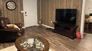 Una sala de estar con un sofá de cuero marrón, una mesa de centro de vidrio, un televisor de pantalla plana sobre un soporte de madera y una puerta con un reloj de pared.