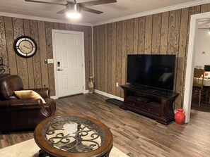 Ein Wohnzimmer mit braunem Ledersofa, einem Couchtisch mit Glasplatte, einem Flachbildfernseher auf einem Holzständer und einer Tür mit Wanduhr.