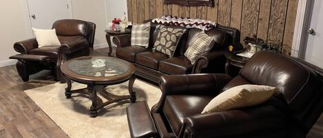 A living room with leather furniture, a glass coffee table, and a wooden wall.